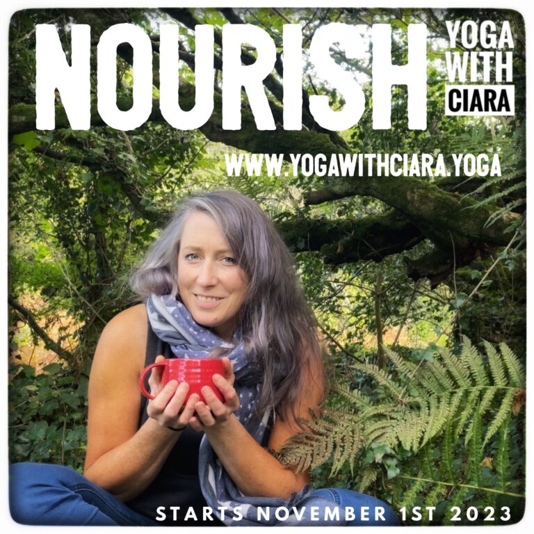 Ciara, with beautiful gray hair, sits serenely under a tree surrounded by lush ferns, her long hair flowing down. She wears a scarf and holds a bright red mug cupped in her hands, radiating a sense of tranquility and warmth in the natural setting. come have a Nourishing November with Ciara
